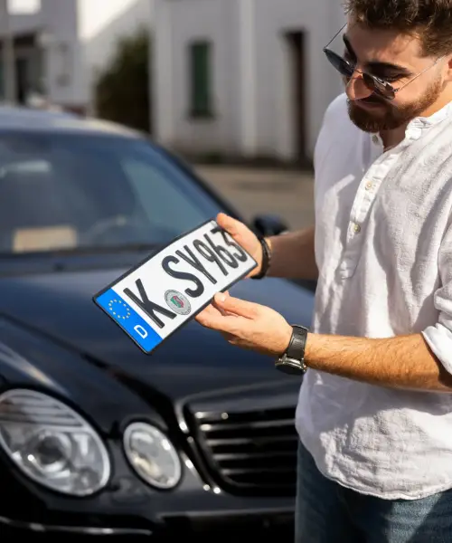 Mann hält ein frisch geprägtes Kölner Kennzeichen mit Zulassungsplakette vor einem schwarzen Auto